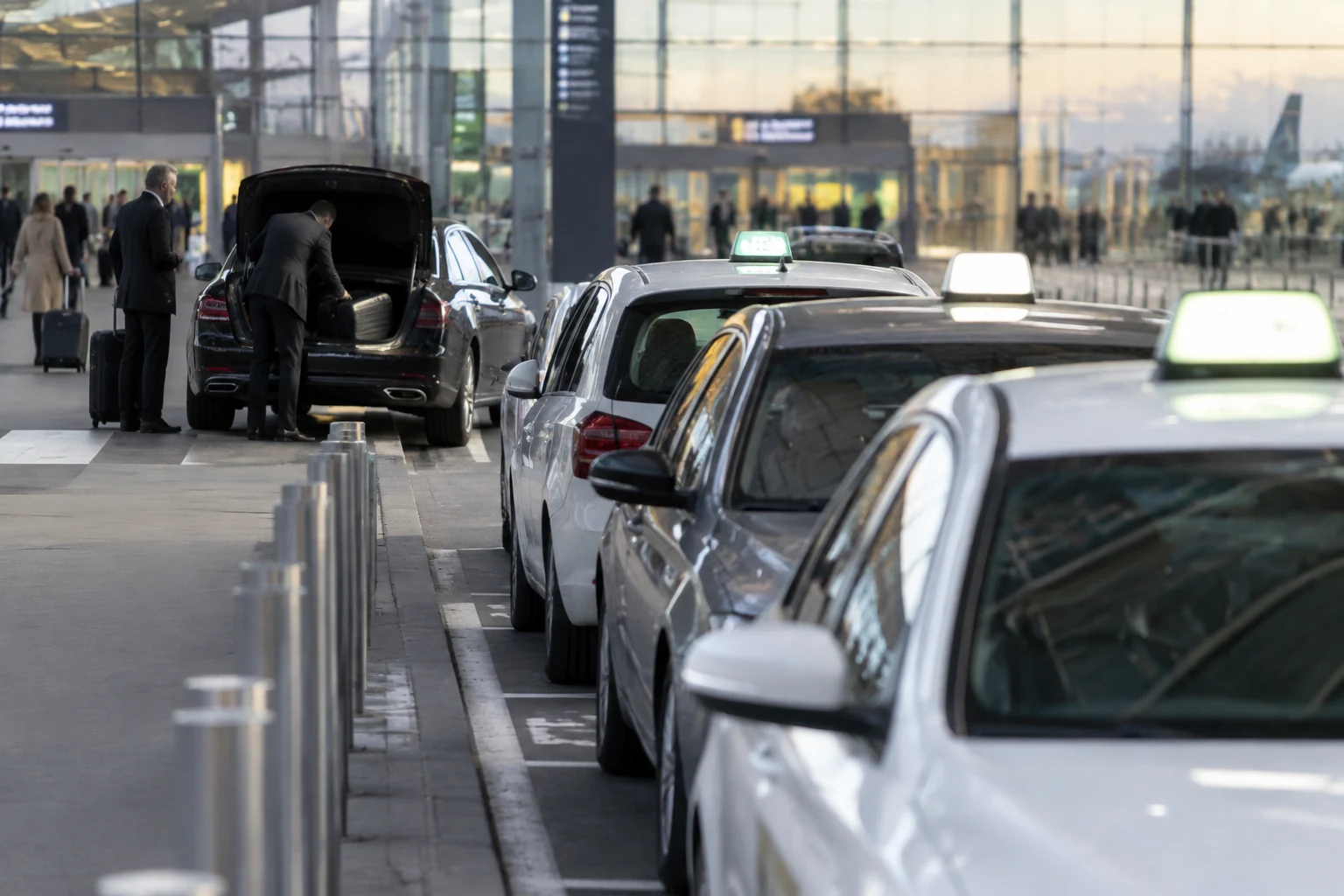 Station de taxis à lumineux devant un terminal d'aéroport, avec une berline VTC noire chargeant les bagages d'un client en arrière-plan
